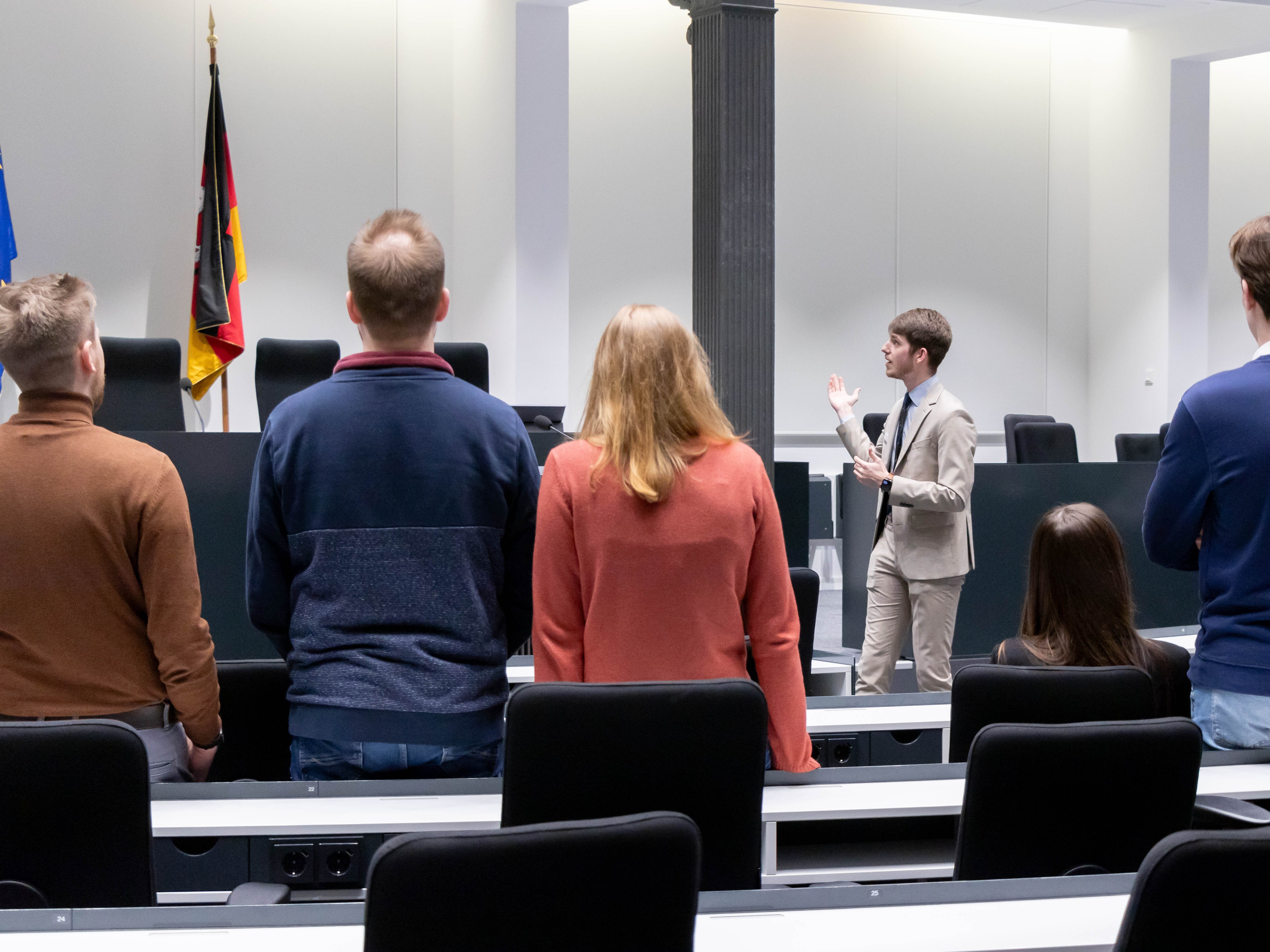 Mehrere Besucherinnen und Besucher stehen mit dem Rücken zur Kamera und sehen sich im Forum des Niedersächsischen Landtages um, während ein Landtagslotse neben dem Redepult steht und in den Raum deutet.