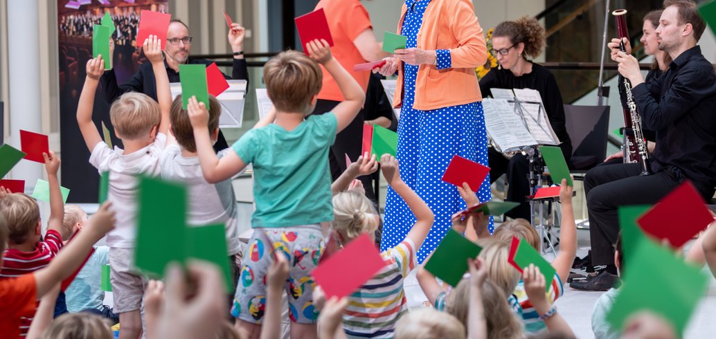 Foto aus der Portikushalle. Im Vordergrund sind viele Kinder zu sehen, die mit dem Rücken zur Kamera auf bunten Kissen auf dem Boden sitzen. Ihnen gegenüber stehen Susanne Grünig sowie die Musikerinnen und Musiker der NDR Radiophilharmonie mit Instrumenten und Notenständern.