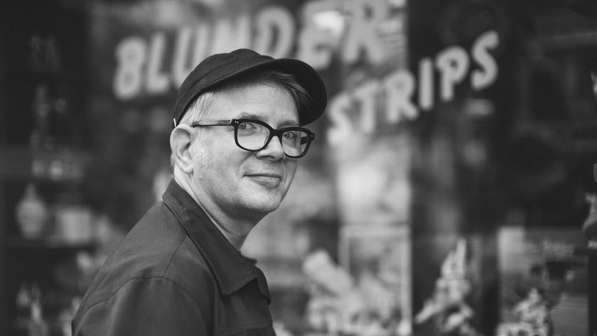 Portraitfoto von Tobi Dahmen in Schwarz-Weiß. Er hat helle Haare, trägt eine Brille mit dunklem Gestell, eine dunkle Kappe und eine dunkle Jacke. Er steht seitlich zur Kamera und dreht den Kopf mit einem leichten Lächeln zum Betrachter. Im Hintergrund ist eine große Schaufensterfront zu erkennen.