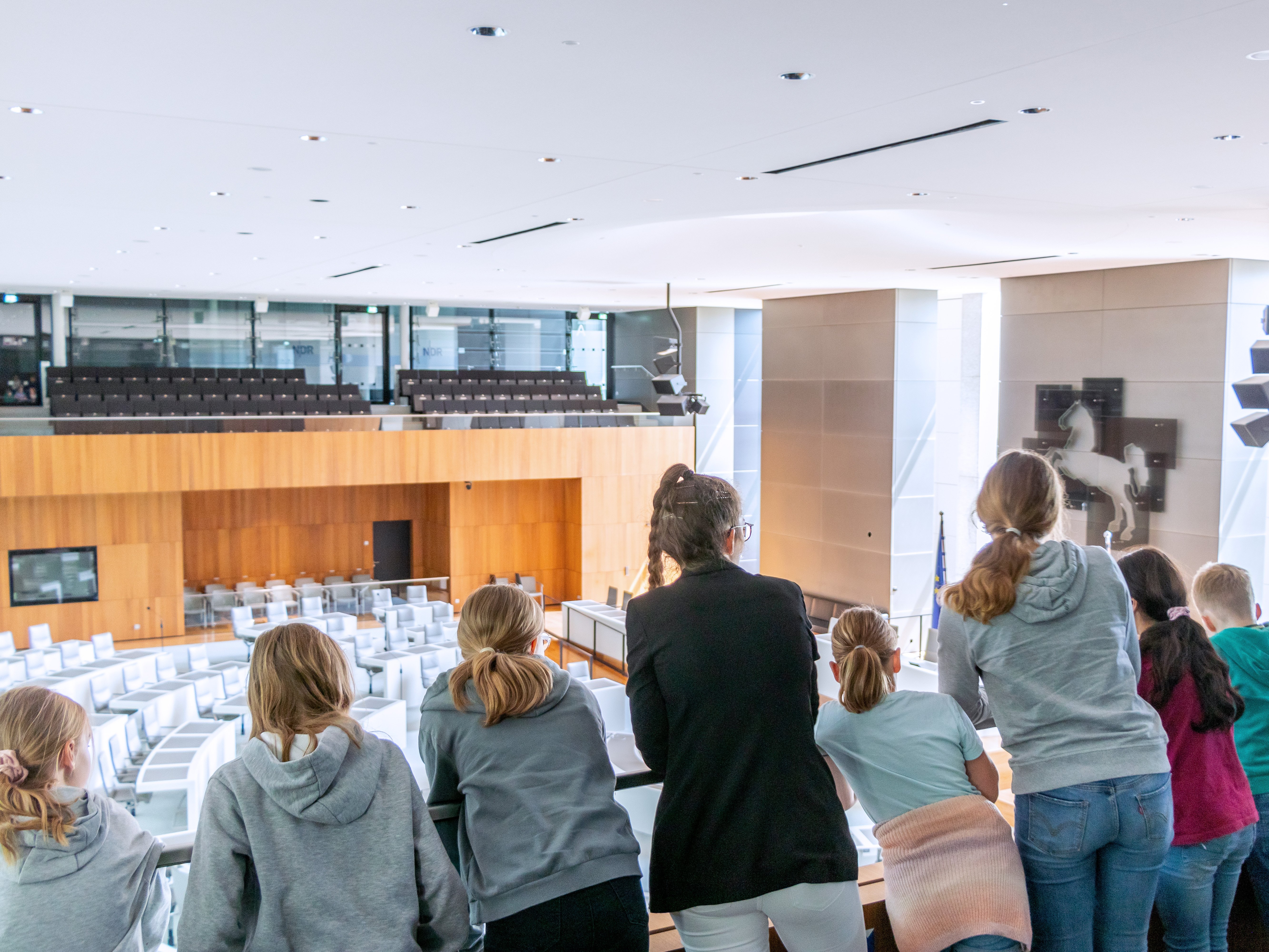 Eine Landtagslotsin umringt von Kindern schaut mit ihnen von der Besuchertribüne hinunter in den leeren Plenarsaal. 