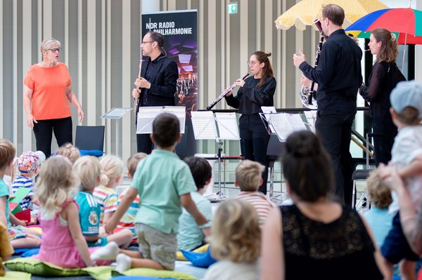 Foto aus der Portikushalle. Im Vordergrund sind viele Kinder zu sehen, die mit dem Rücken zur Kamera auf bunten Kissen auf dem Boden sitzen. Ihnen gegenüber stehen Susanne Grünig sowie die Musikerinnen und Musiker der NDR Radiophilharmonie mit Instrumenten und Notenständern.